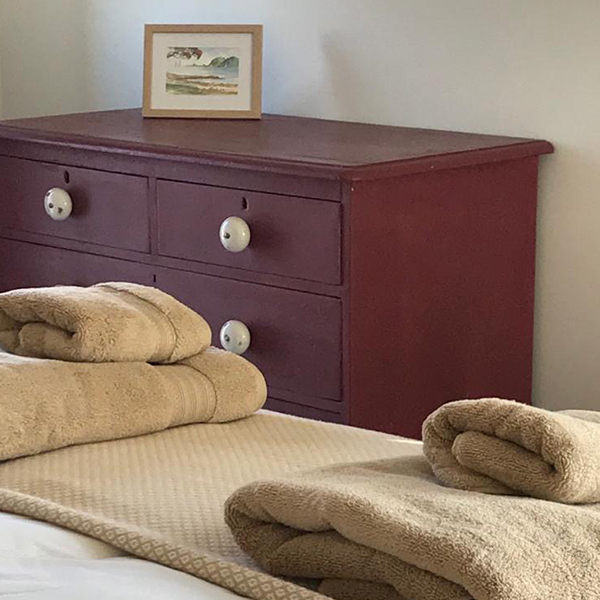 Corner of a bedroom with a red chest of drawers with towels sitting on the end of the double bed